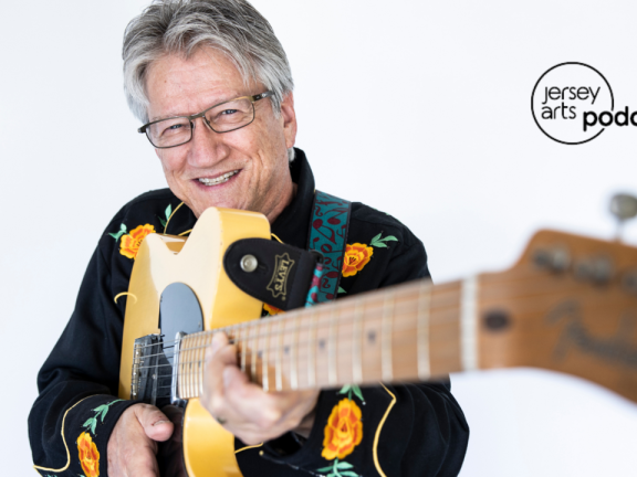 Richie Furay holding a guitar towards the camera with the Jersey Arts Podcast logo on top