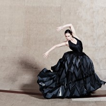 "Female dancer in a flowing black gown striking an expressive pose against a curved wooden backdrop.