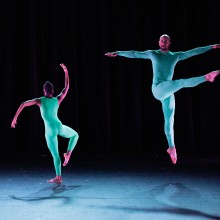 Two dancers on stage dressed in green unitards - one in the air with one leg stretched forward; the other in an angular shap