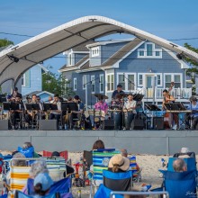 Big band performs under a white canopy on a beach stage while an audience in beach chairs watches; saxophones, drums, uprigh