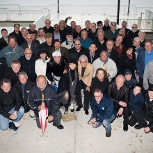 Large group portrait of about 60 adults posing on a waterfront dock, wearing jackets and hats, smiling up at the camera with