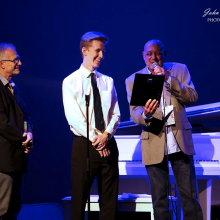 Three people onstage under blue lights; a man reads from a plaque into a microphone while a smiling young man in a shirt and