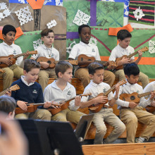 Elementary school students seated on risers play ukuleles during a school music performance, wearing matching white uniforms