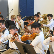 Students in a school orchestra play violins, brass, and woodwind instruments during a rehearsal or concert, seated behind mu