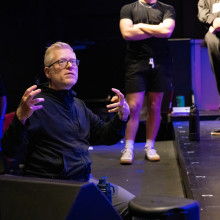Artistic director seated at the edge of a stage gesturing while speaking to actors during a rehearsal, with performers stand