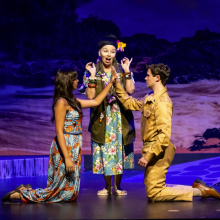 Three performers on stage in a scene from South Pacific, with one actor standing between two kneeling characters against a t