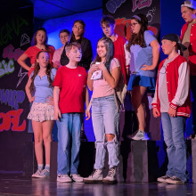 Teens perform a contemporary musical number on stage under blue lighting, with a central girl holding a notebook as the grou