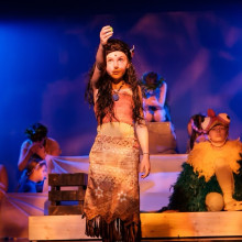 Young performer in an island-inspired costume stands center stage holding a glowing object, with other children seated behin
