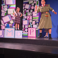 Actor portraying a strict headmistress gestures on stage beside a young girl in a school uniform, set against a colorful wal