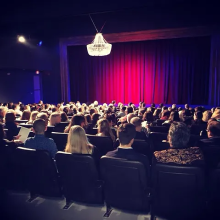 A crowd of people sitting in chairs at an auditorium with a red curtain pulled onstage