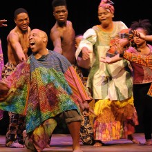 A group of performers onstage dance around a man on his knees singing, all wearing traditional African garments