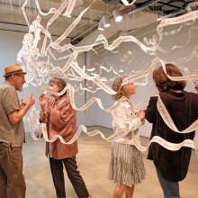 four people stand in a gallery talking amongst the artwork: long strips of paper with writing on it strung around the room