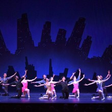 A group of dancers on stage in front of a set design made to resemble a city