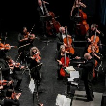 ensemble in all black concert attire performs string instruments in front of conductor