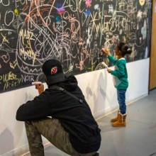 a man and a child both draw on a large chalkboard, covered in art from a collection of other people