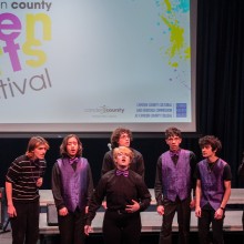 High schoolers in black shirts with purple vests sing in front of a “Teen Arts Festival” sign