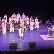 a choir of people wearing all white with black scarves perform on a blue stage