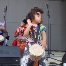 a child in African garments with a drum around their neck performing onstage
