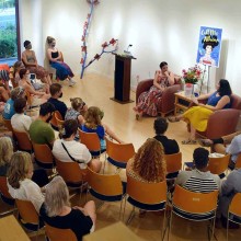 an author sits in front of a crowd in a room with a poster of her book titled “call Me Athena” behind her