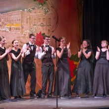 a choir of young girls and boys in fancy black attire clapping and singing on stage, in front of a Christmas themed backdrop