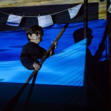A man holding a paddle sitting in a pretend canoe on a stage swathed in blue light