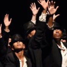Men in a black box theatre wearing suits and black hats reaching up under a spotlight