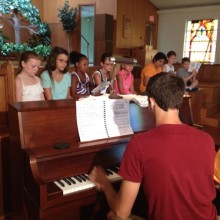 A teacher sits at a piano and goes over the songbook of a musical with a group of young actors