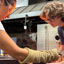 Two young adults helping each other in a glassblowing class