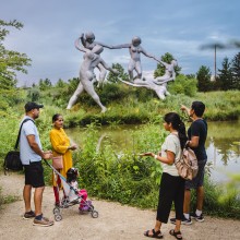 A multigenerational family on the path in front of Daydream, a large silver sculpture of five female figures dancing.