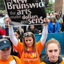 protestors in orange shirts gathered, one person holding a sign that says “In New Brunswick, the arts make dollars and sense”