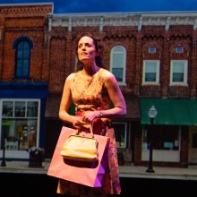 an actress in vintage looking costume holds a purse in front a main street set under a spotlight
