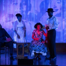 a group of African American actors perform under blue stage lights with an image of Emmett Till and his mother behind them