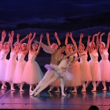 A pas de deux in front of a line of dancers under soft pink stage lights with a blue backdrop