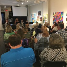 an artist stands at a podium and gives a talk in front of a crowd inside an art gallery