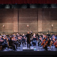 A conductor stands in front of a large orchestra on a stage