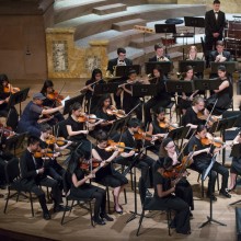 A close-up on a performance of a youth orchestra, the performers wearing all black and following their conductor