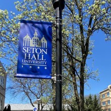 A blue sign for Seton Hall University hung up on a lamppost in from of some trees