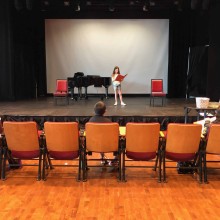 a child stands on stage with music book in hand an performs an audition in front of her theatre teacher and peers