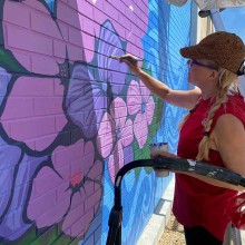 Woman paints purple and pink flowers on a brick wall as part of an outdoor mural