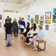 amidst standing viewers, three woman sit on a bench in an art gallery and look at the artwork