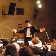 an image of a man in a suit with a bowtie attire conducting a group of musicians