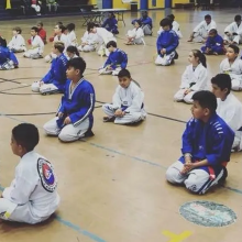 kids in white and blue robes sit on a gym floor during a karate class