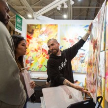 A man displays a large canvas painting to two viewers in an art fair booth