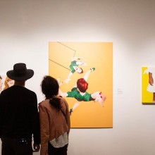 A couple stands in front of three paintings hung on a gallery wall