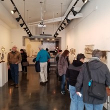 about 8 people look around an art gallery, a long hall with lighting fixtures above various fiber works