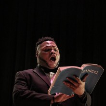 soloist singing under stage lights holding a Handel music book