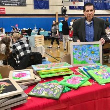 An older man in a sports coat showing off a series of floral paintings at an art fair in a gymnasium