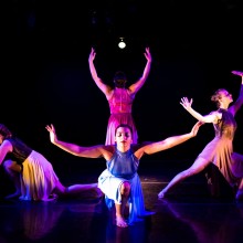 four women pose in a dance movement on a dark stage under a spotlight