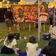 An artist spray paints a graffiti style sign that says “Arts After Dark” while children look on