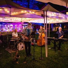 A band performs at night outdoors in the grass under a tent covered in string lights